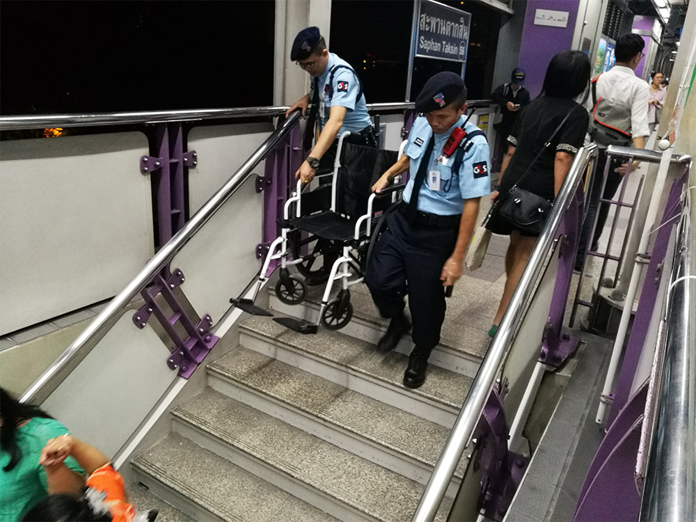 Private security carry a commuter's wheelchair down the stairs Tuesday at BTS Saphan Taksin. Three years after the highest court gave the city 12 months to make all stations wheelchair-accessible, the work remains incomplete.