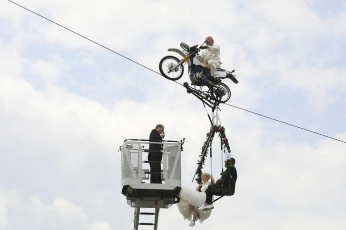 Pastor Stefan Gierung, at left, stands in a cage atop of a fire service ladder in front of bride Nicole Backhaus, center, and groom Jens Knorr, right, both sitting in a swing dangling under a motorcycle with artist Falko Traber, top, during the wedding ceremony atop a tightrope Saturday in Stassfurt, Germany. Photo: Peter Gercke/ DPA Pastor Stefan Gierung, at left, stands in a cage atop of a fire service ladder in front of bride Nicole Backhaus, center, and groom Jens Knorr, right, both sitting in a swing dangling under a motorcycle with artist Falko Traber, top, during the wedding ceremony atop a tightrope Saturday in Stassfurt, Germany. Photo: Peter Gercke/ DPA