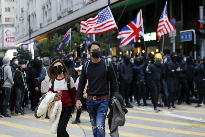 A masked couple walk by pro-democracy protesters who wave US and British flags as they gather on a street in Hong Kong, Sunday, Dec. 8, 2019. (AP Photo/Vincent Thian)