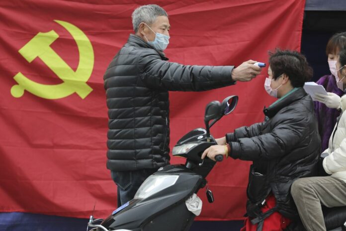 A volunteer stands in front of a Communist Party flag as he takes the temperature of a scooter driver at a roadside checkpoint in Hangzhou in eastern China's Zhejiang Province, Monday, Feb. 3, 2020. (Chinatopix via AP)