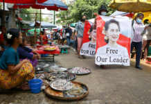 Myanmar Puts Suu Kyi on Trial on Charges Critics Call Bogus Anti-coup protesters walk through a market with images of ousted Myanmar leader Aung San Suu Kyi at Kamayut township in Yangon, Myanmar Thursday, April 8, 2021. Photo: AP
