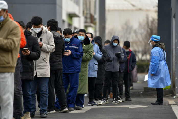 In this photo released by Xinhua News Agency, a worker wearing a protection suit looks at residents wearing face masks to protect from the coronavirus as they line up for the coronavirus test during a mass testing in north China's Tianjin municipality, Sunday, Jan. 9, 2022. Photo: Zhao Zishuo / Xinhua via AP