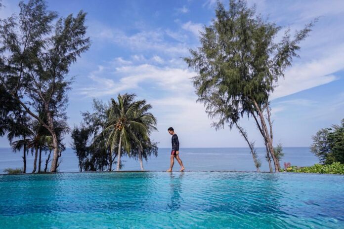 thailand-beach-resort-pool A man walking along an infinity pool by the beach surrounded by palm trees at a Thailand beach resort.