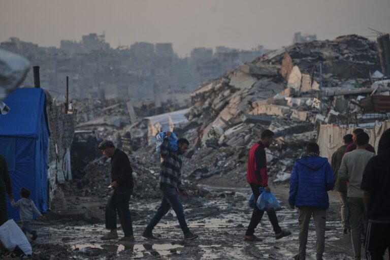 a photograph of people walking in front of a large background of rubble with murky skies in gaza