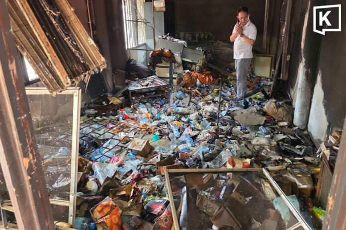 A room several meteres long and a few metres wide is strewn with trash. Windows let in daylight and there is an official in a whiteshirt standing in the garbage, which covers his ankles.