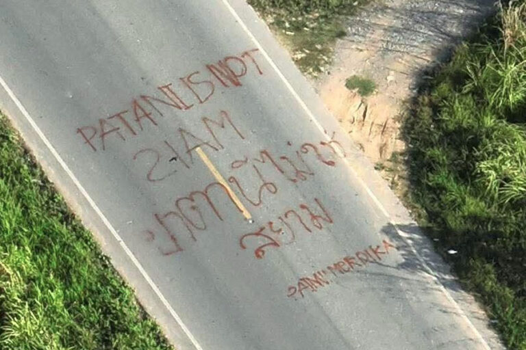 An overhead photo of a highway where hastily-written graffiti says "PATANI IS NOT SIAM" and ปัตตานีไม่ใช่สยาม. The "S" in "SIAM" is drawn backwards. There are some words beneath in the same red font, but are difficult to make out.