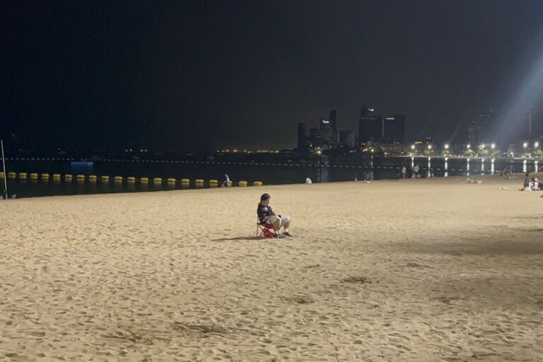 a man sitting in a beach chair faces away from the water at Pattaya Beach, late at night. The beach is lit up with nearby streetlights.